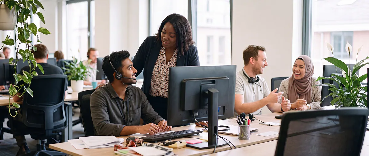 A team lead leaning over a colleague's screen in a busy service centre